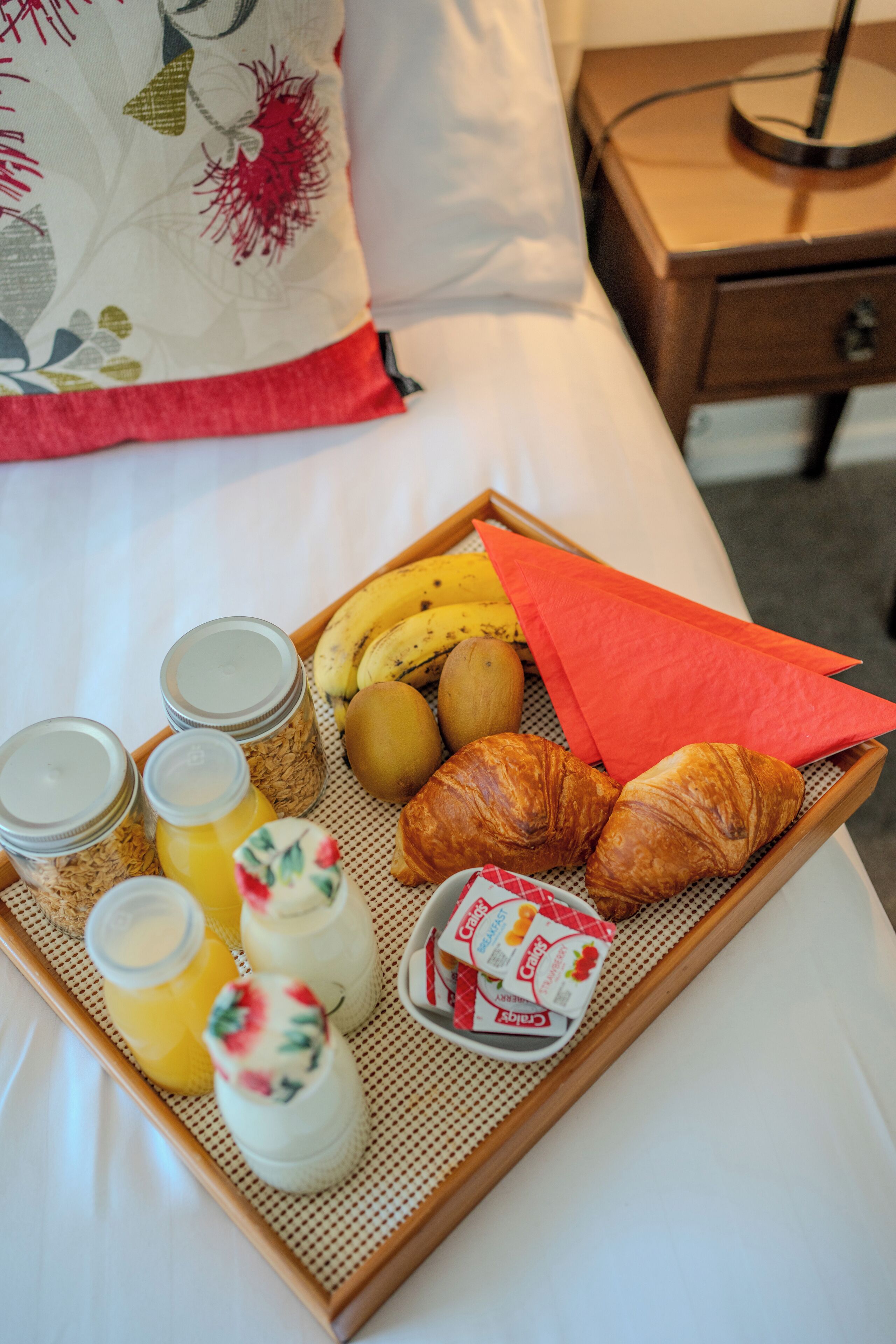 Ontbijtplateau met croissants, bananen, kiwi, ontbijtgranen, yoghurt en sap op een bed.