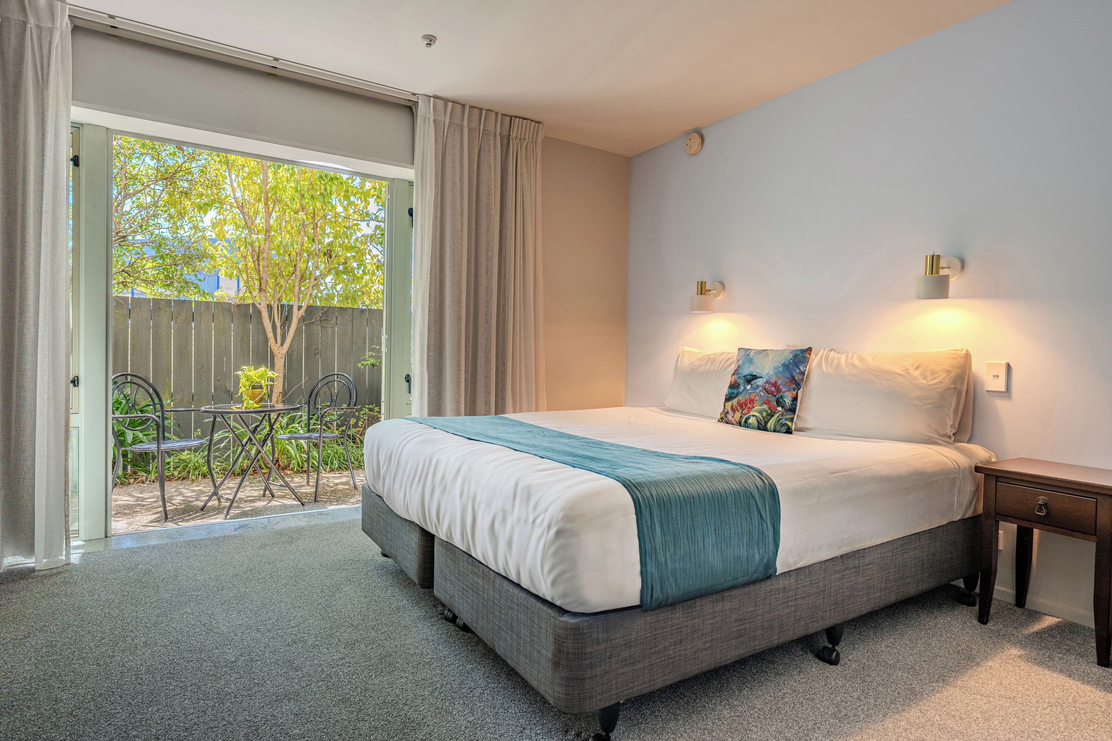 A bedroom with a bed, a nightstand, a window with curtains, and a view of a patio with a table and chairs.