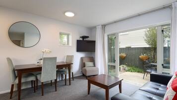 A room with a dining table, chairs, a sofa, a TV mounted on the wall, and a sliding glass door leading to an outdoor area with a garden.