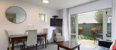 A room with a dining table, chairs, a sofa, a TV mounted on the wall, and a sliding glass door leading to an outdoor area with a garden.