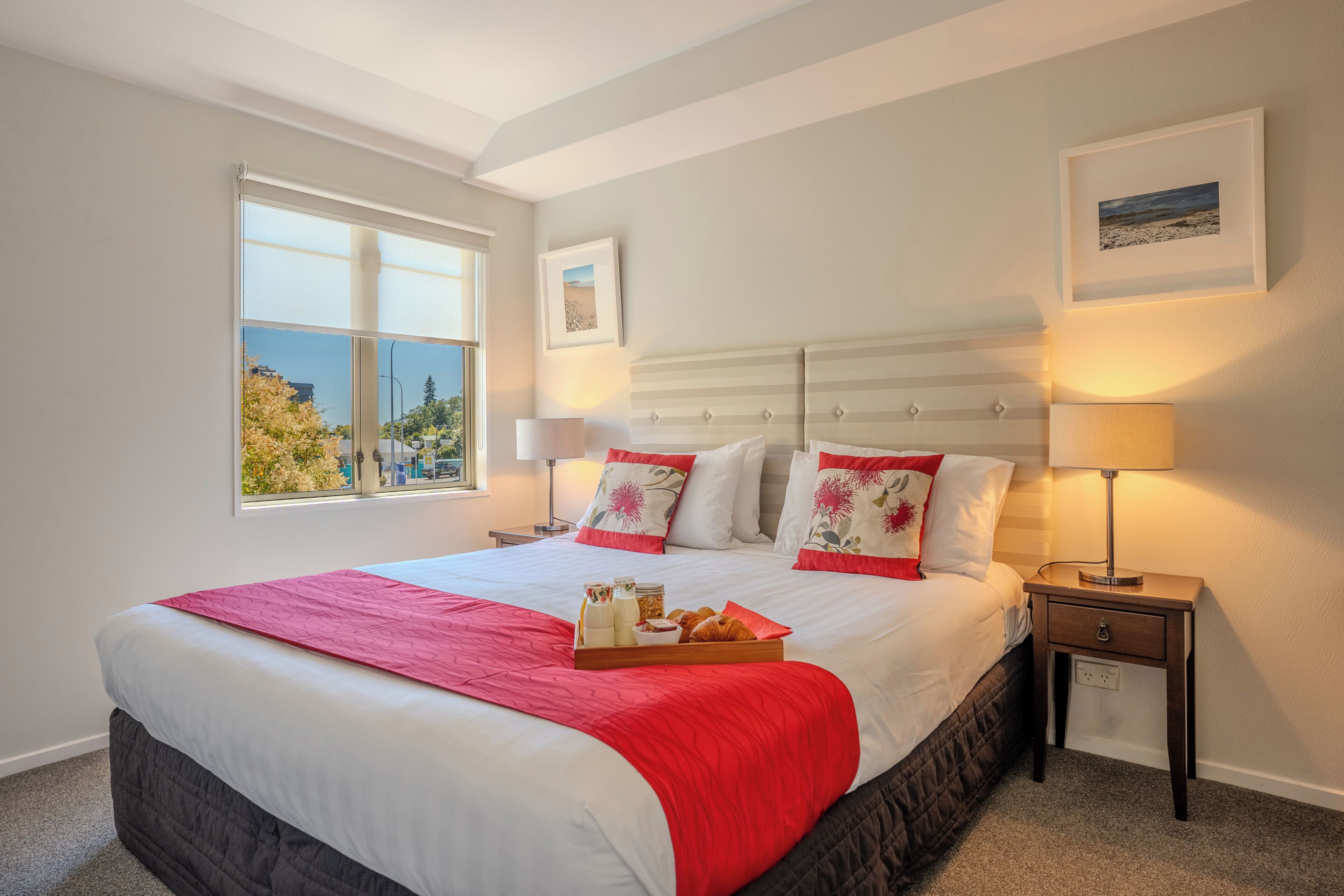 A neatly arranged bedroom with a large bed, bedside lamps, a window with a view, and a tray of breakfast items.