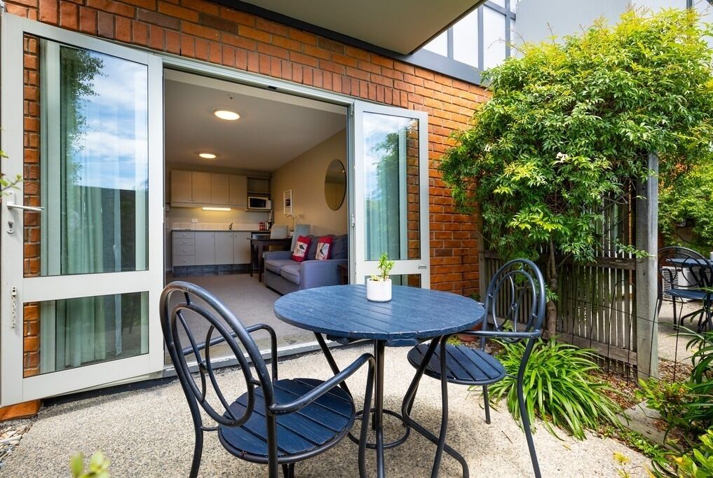 An open patio with a round table and chairs, a sliding glass door leading to a kitchen, and a brick wall.