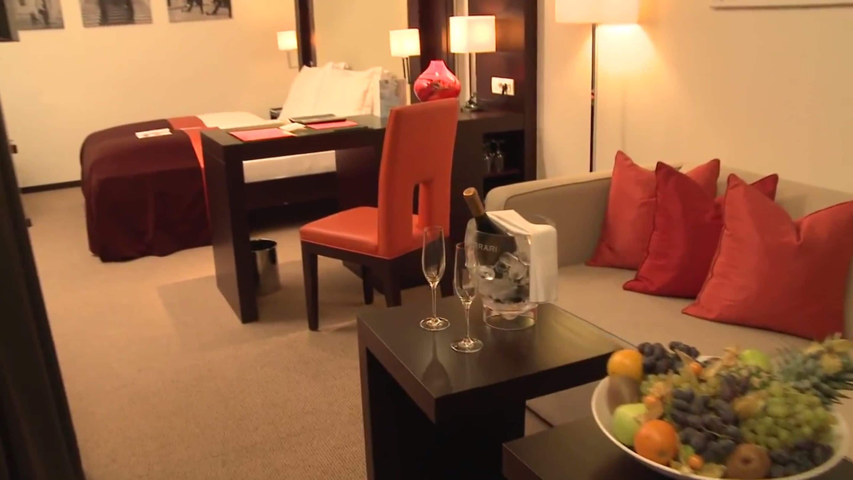 Habitación de hotel con una mesa de comedor, una silla roja, un sofá con cojines rojos, un frutero y una minibar.