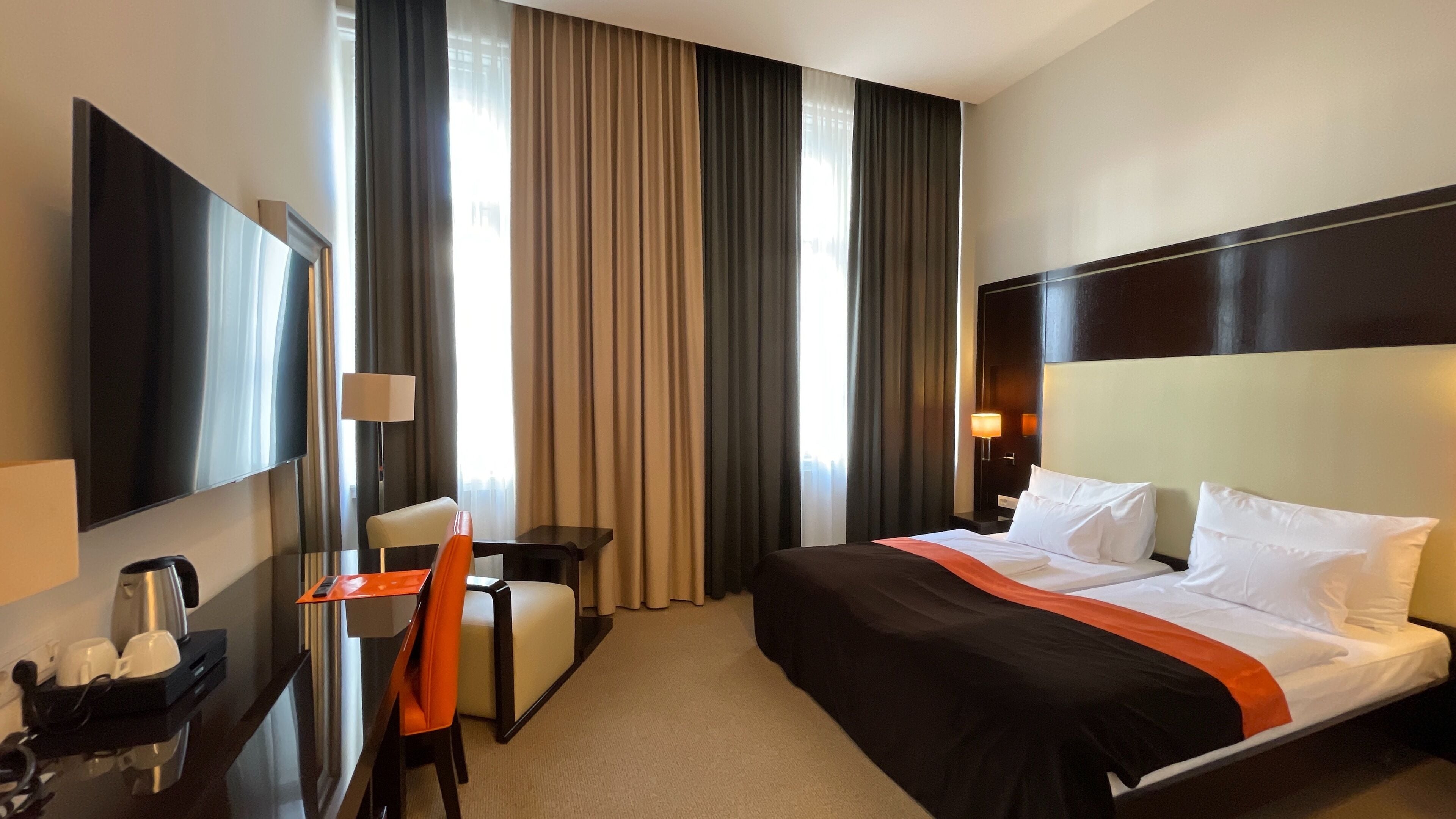 Habitación de hotel con cama, escritorio, silla, televisor y ventana con cortinas.