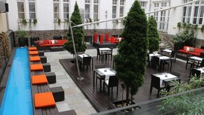 Een buitenterras met zitplaatsen en tafels, een zwembad en een gebouw met ramen op de achtergrond.