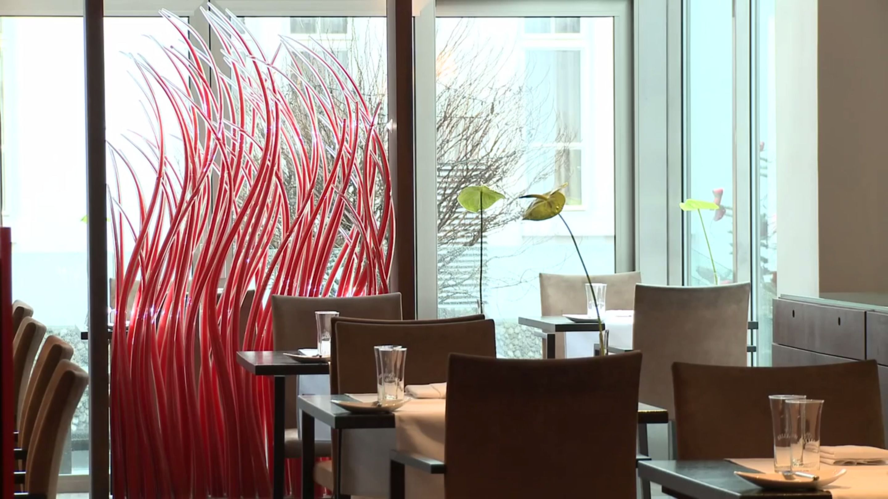 A modern dining area with tables and chairs, large windows, and decorative red elements.