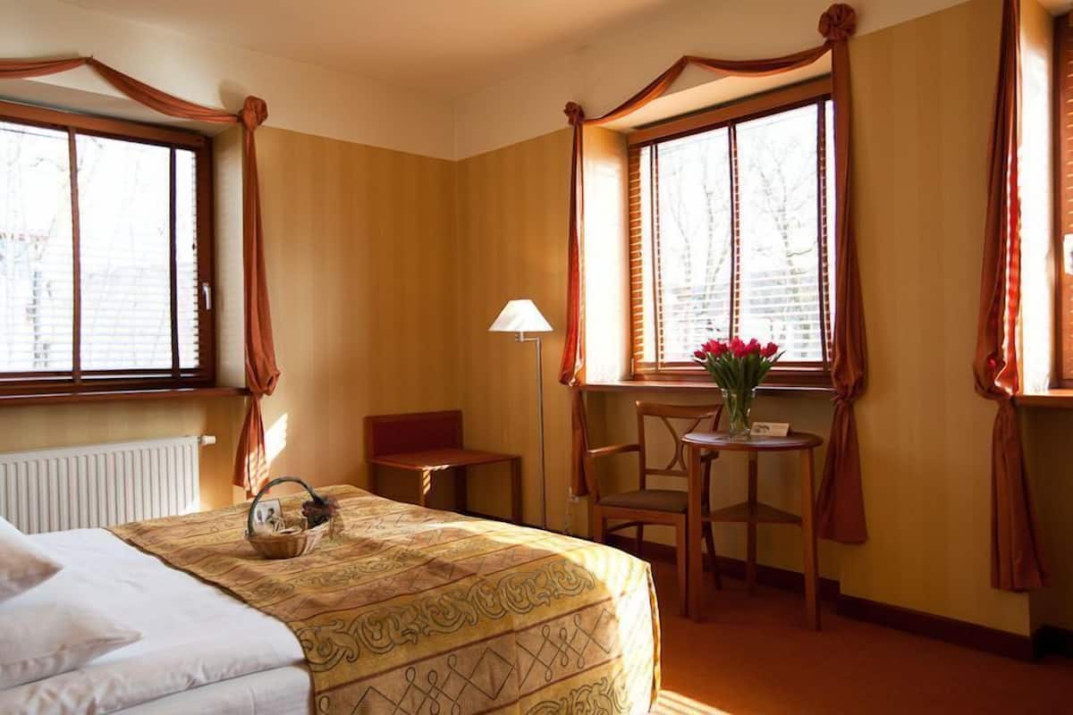 Habitación de hotel con una cama, una mesita con lámpara, una silla y un ventanal con cortinas.