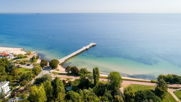 Vista aérea de una zona costera con un muelle que se adentra en el mar, rodeada de vegetación y un pequeño asentamiento con edificios y una carretera.