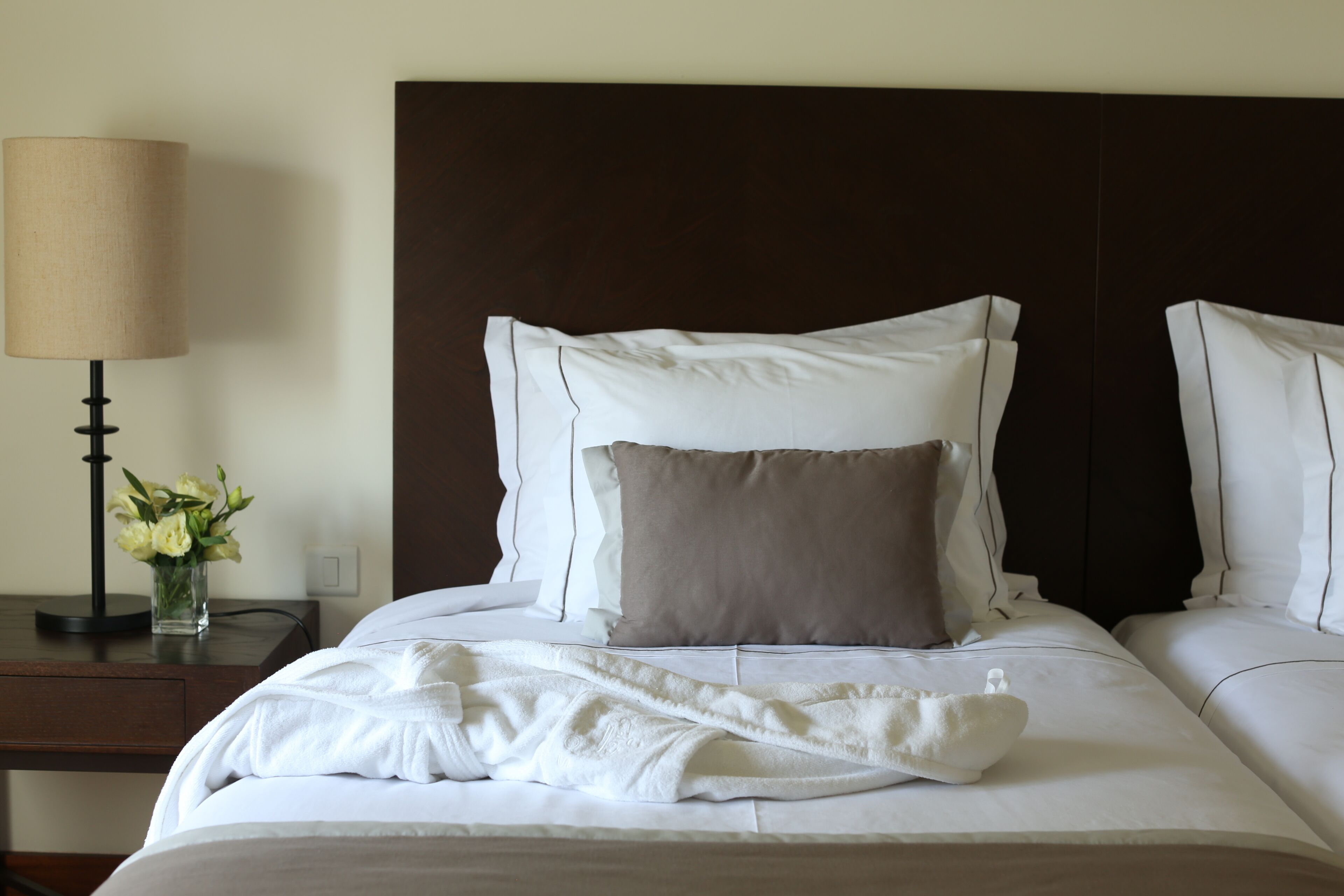 A neatly made bed with white linens and two pillows, a bedside table with a lamp and a vase of flowers, and a dark headboard.