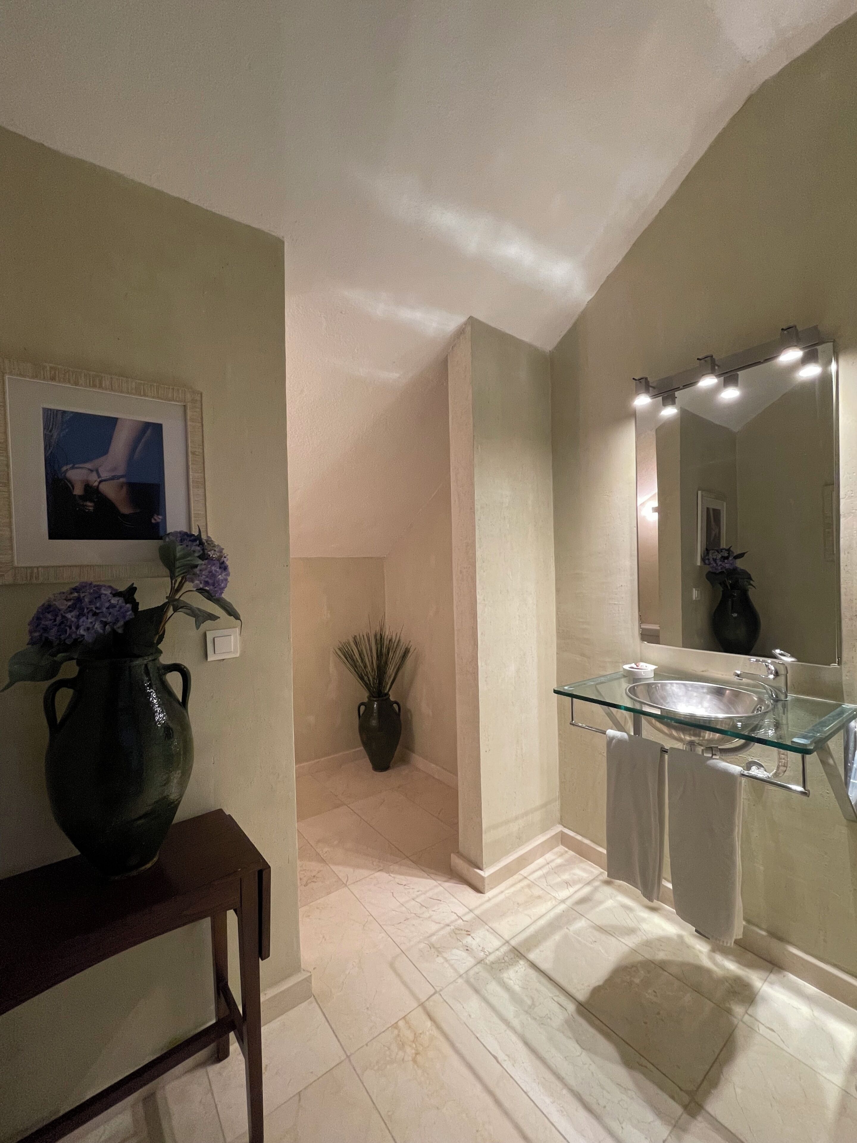 Un baño con un lavabo de cristal, un espejo con luces, un florero con flores y un cuadro en la pared.