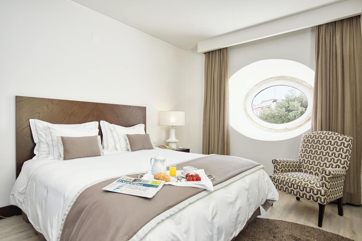 A neatly made bed with a breakfast tray, a chair, and a window with curtains.