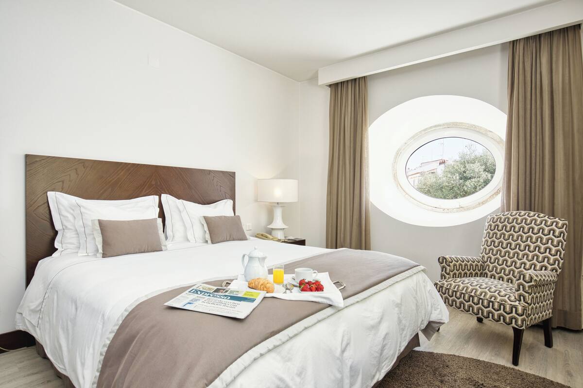 A neatly made bed with a breakfast tray, a chair, and a window with curtains.