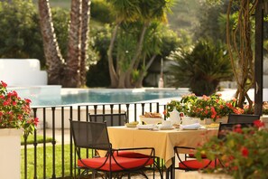 An outdoor dining area with a table set for two, surrounded by chairs with red cushions, a pool in the background, and lush greenery. - Pateo dos Solares Charm Hotel (Estremoz)