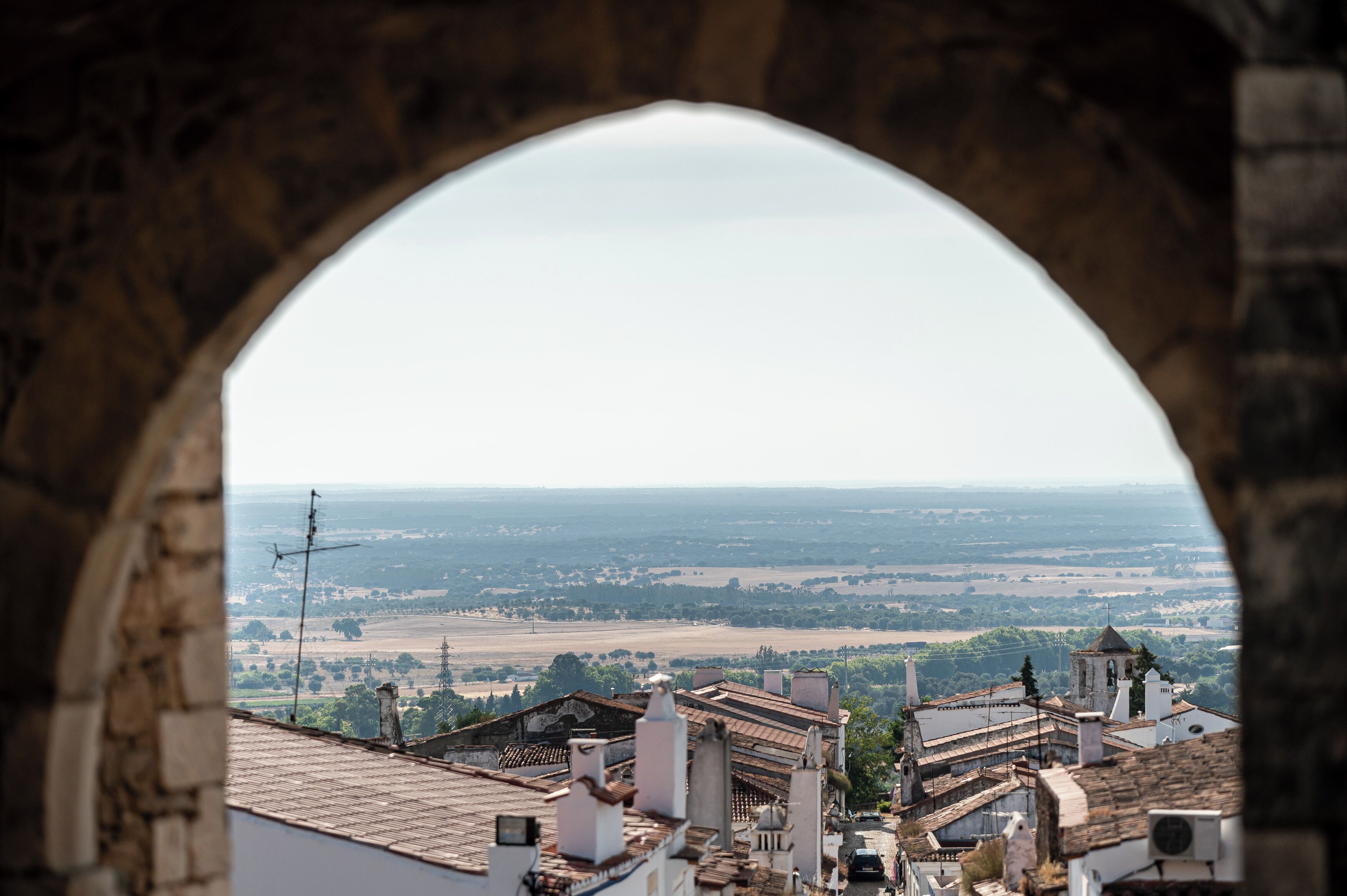 Vista a través de un ventanal de arco que da a un pueblo con edificios históricos y un paisaje lejano.
