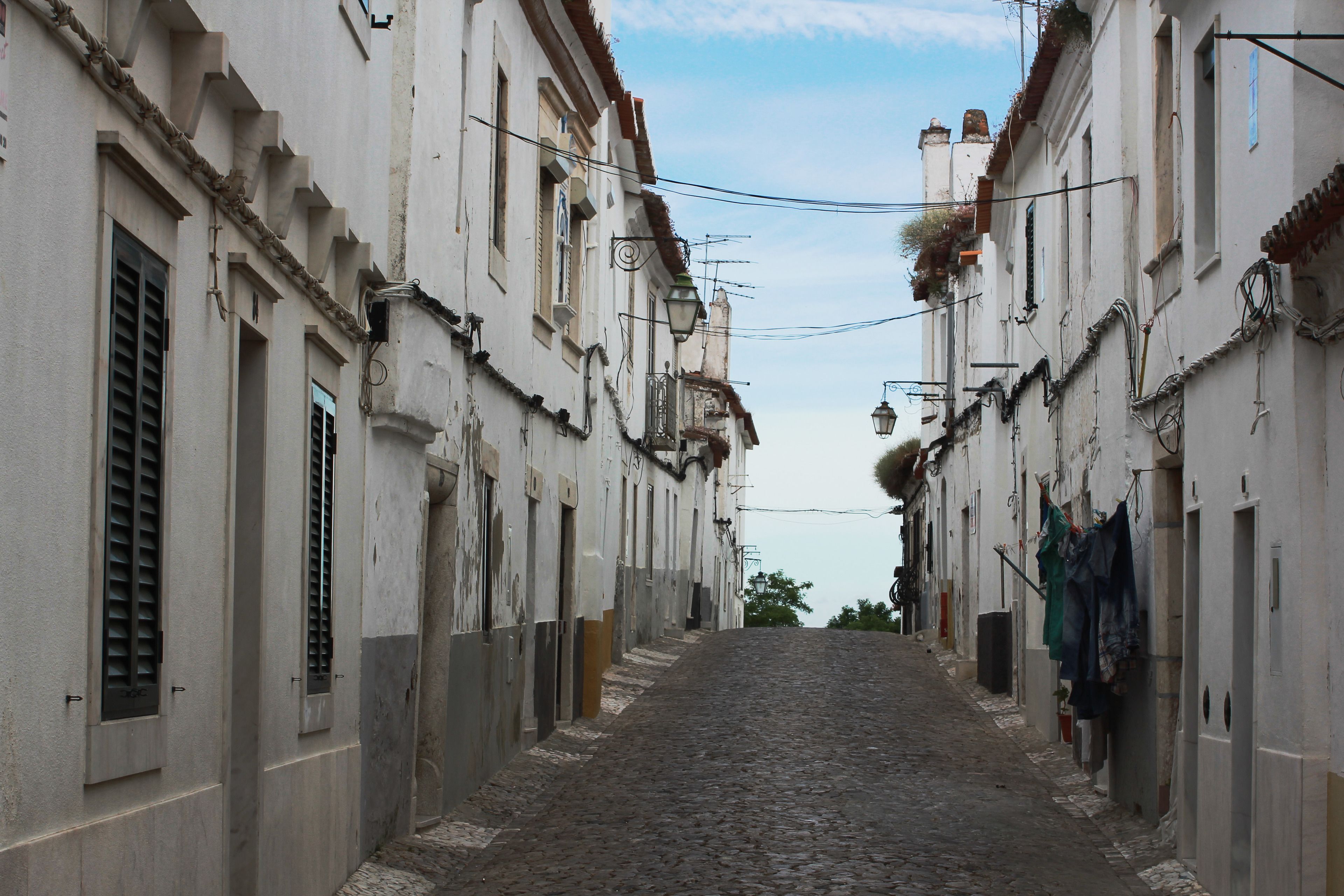 Uma rua de paralelepípedos estreita, ladeada por edifícios históricos com janelas protegidas por persianas e varandas.