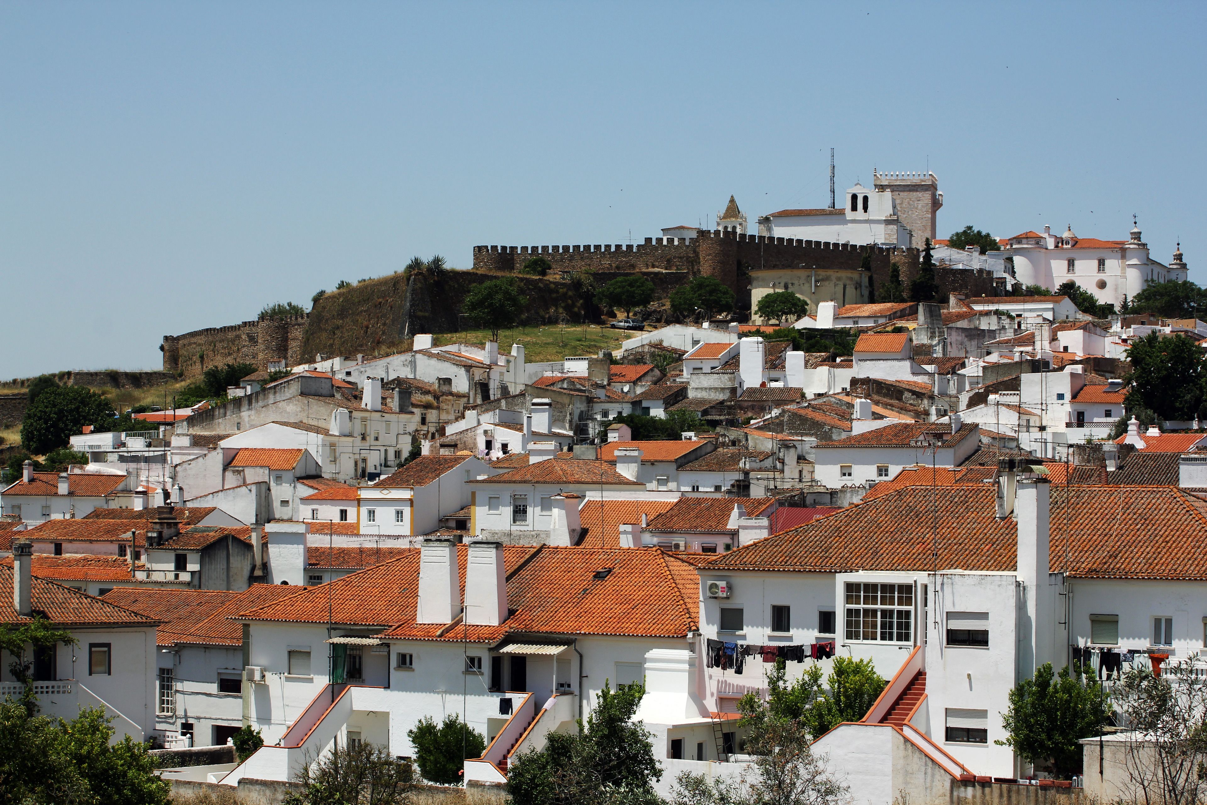 A historic town with white buildings and red-tiled roofs, a prominent castle on a hill, and a clear blue sky.