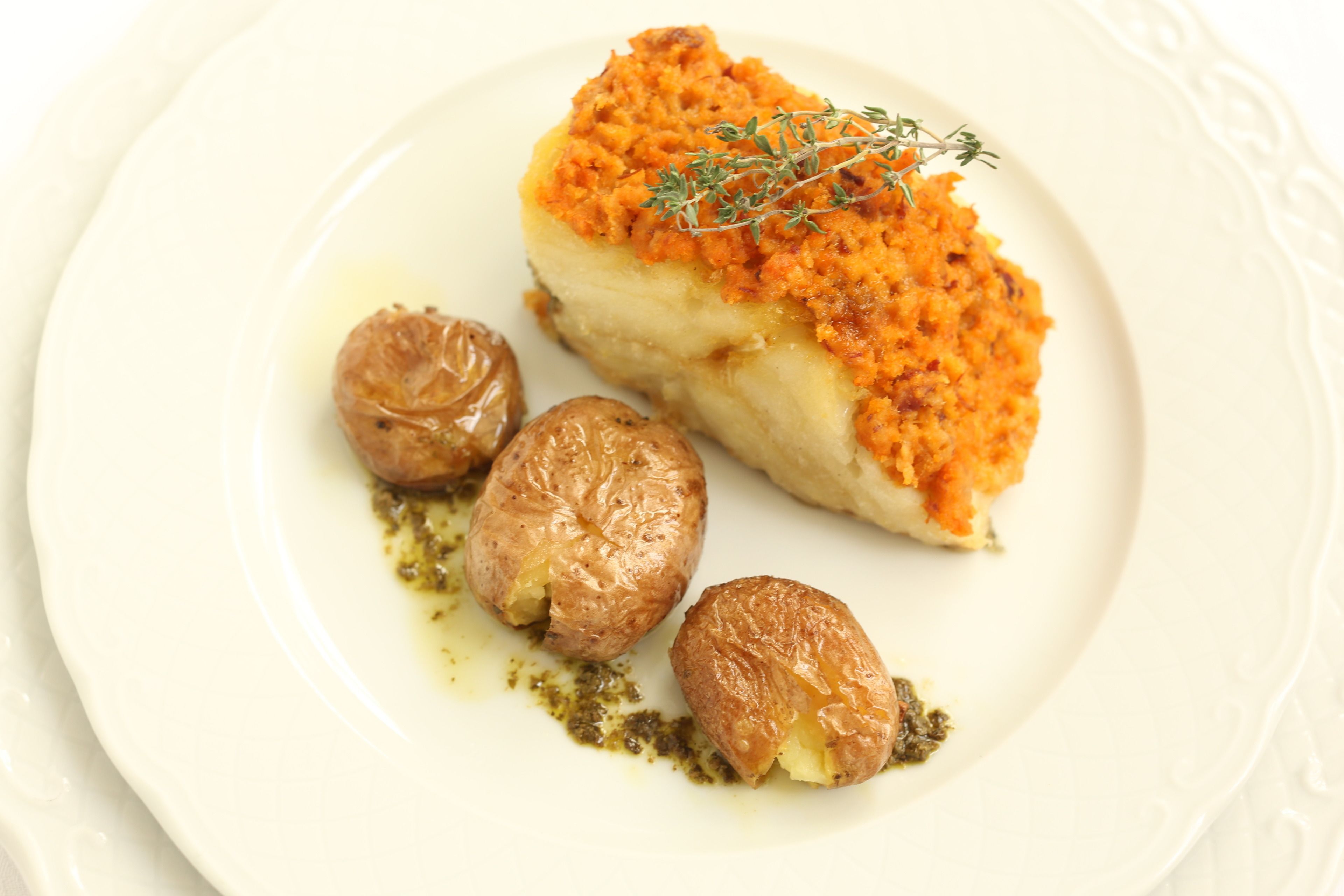 A plate with a piece of fish topped with a golden-brown crust, accompanied by two roasted potatoes and a sprig of thyme.