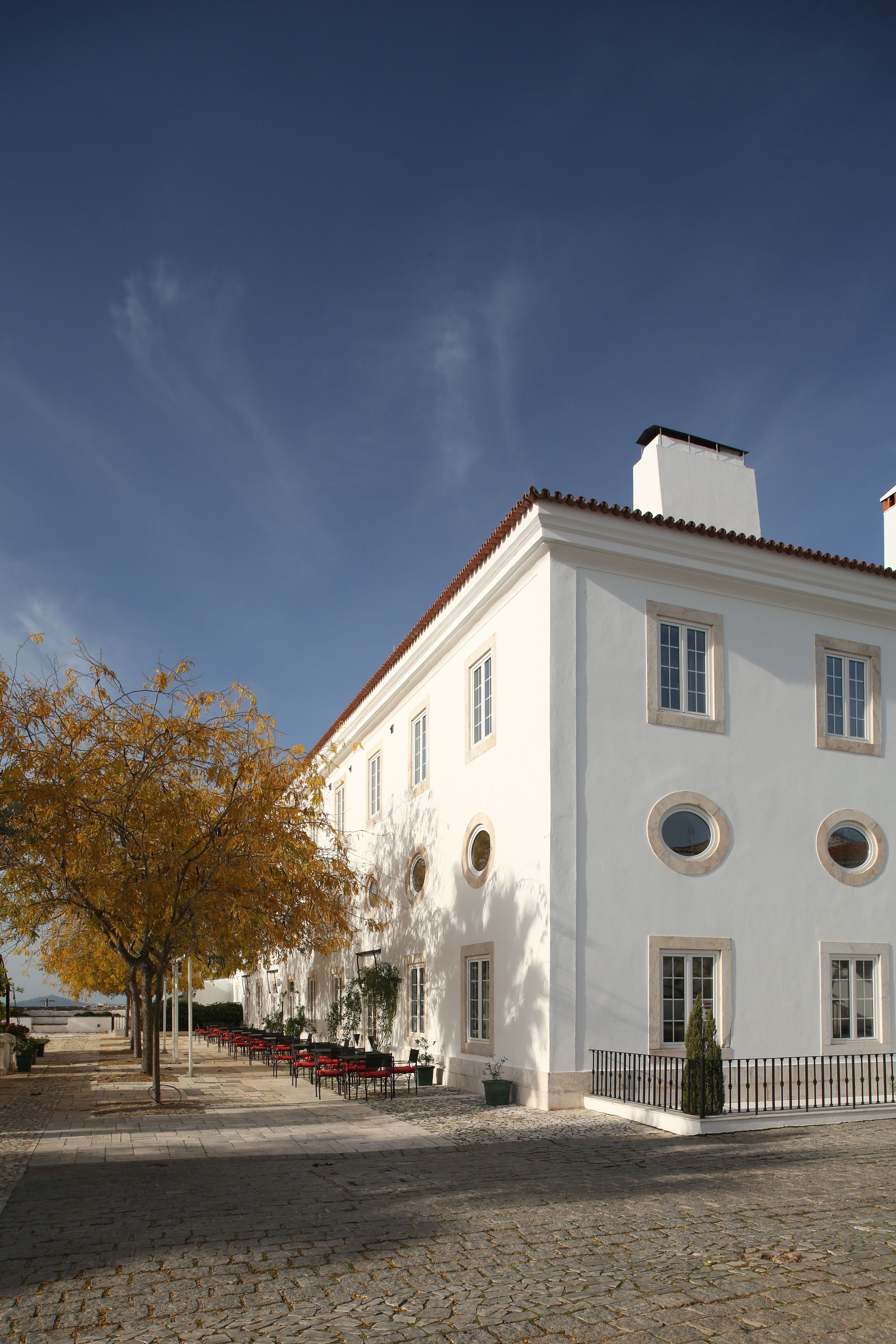 A white building with a red-tiled roof, multiple windows, and a paved courtyard with outdoor seating.