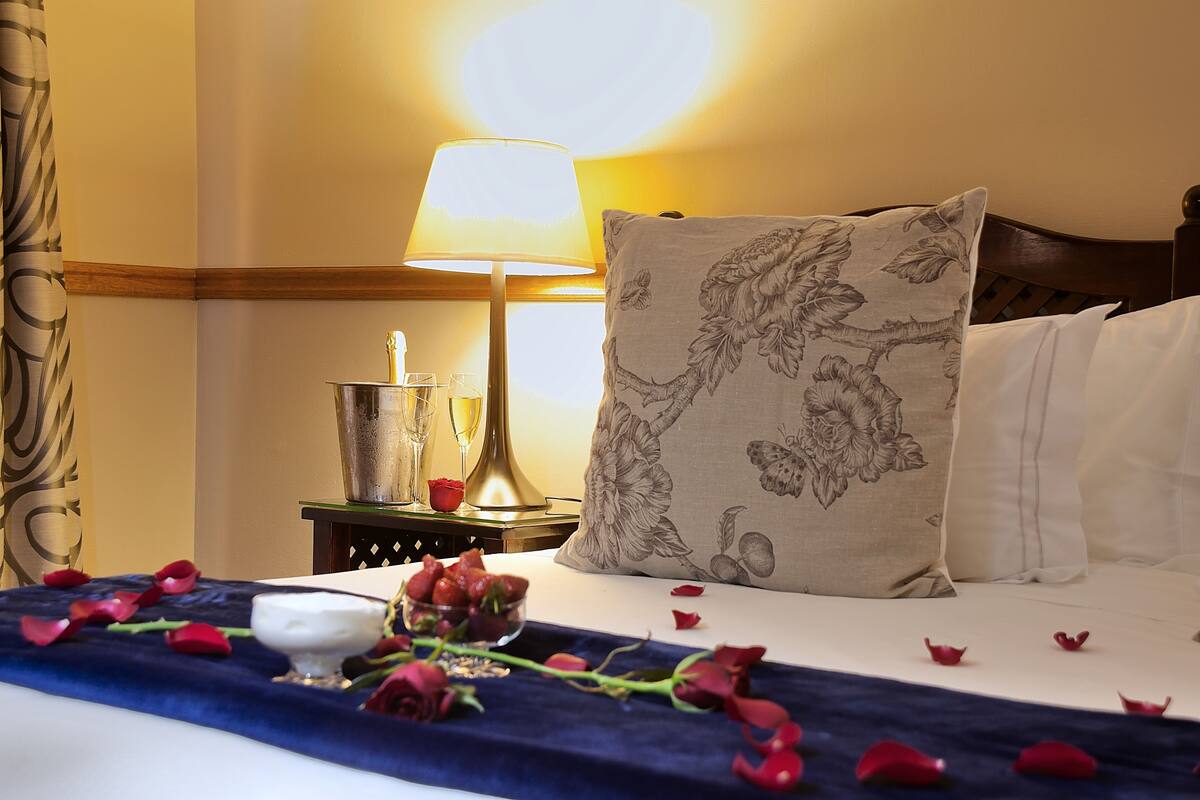 Een opgemaakt bed met een bloemenkussen, een nachtkastje met een lamp en een vaas met rozen.