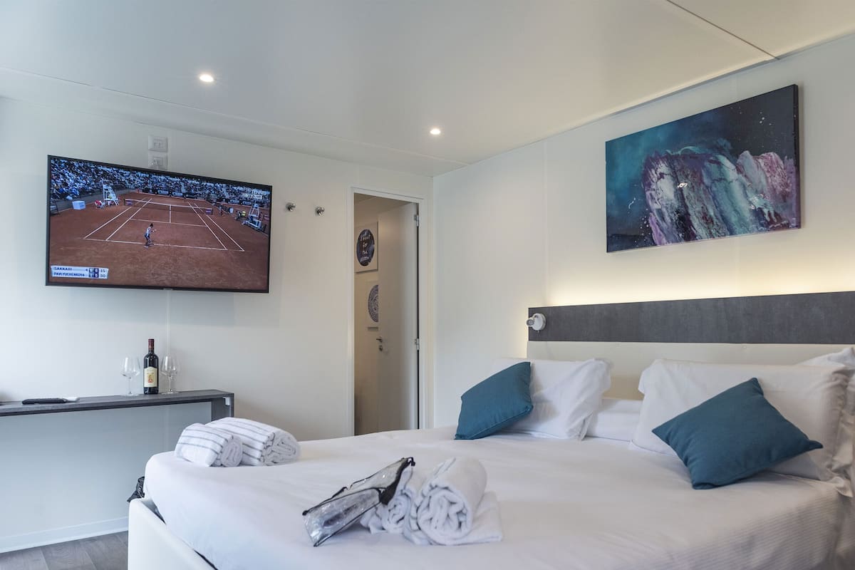 A hotel room with a bed, a TV displaying a tennis match, a painting, and a bottle of wine on a side table.
