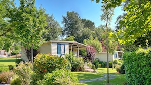A single-story mobile home with a covered porch, surrounded by well-maintained gardens and trees. - Flaminio Village Bungalow Park - Campground (Rome)