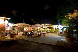 Un área de comedor al aire libre con mesas y sillas, rodeada de plantas en macetas e iluminada por luces colgantes.