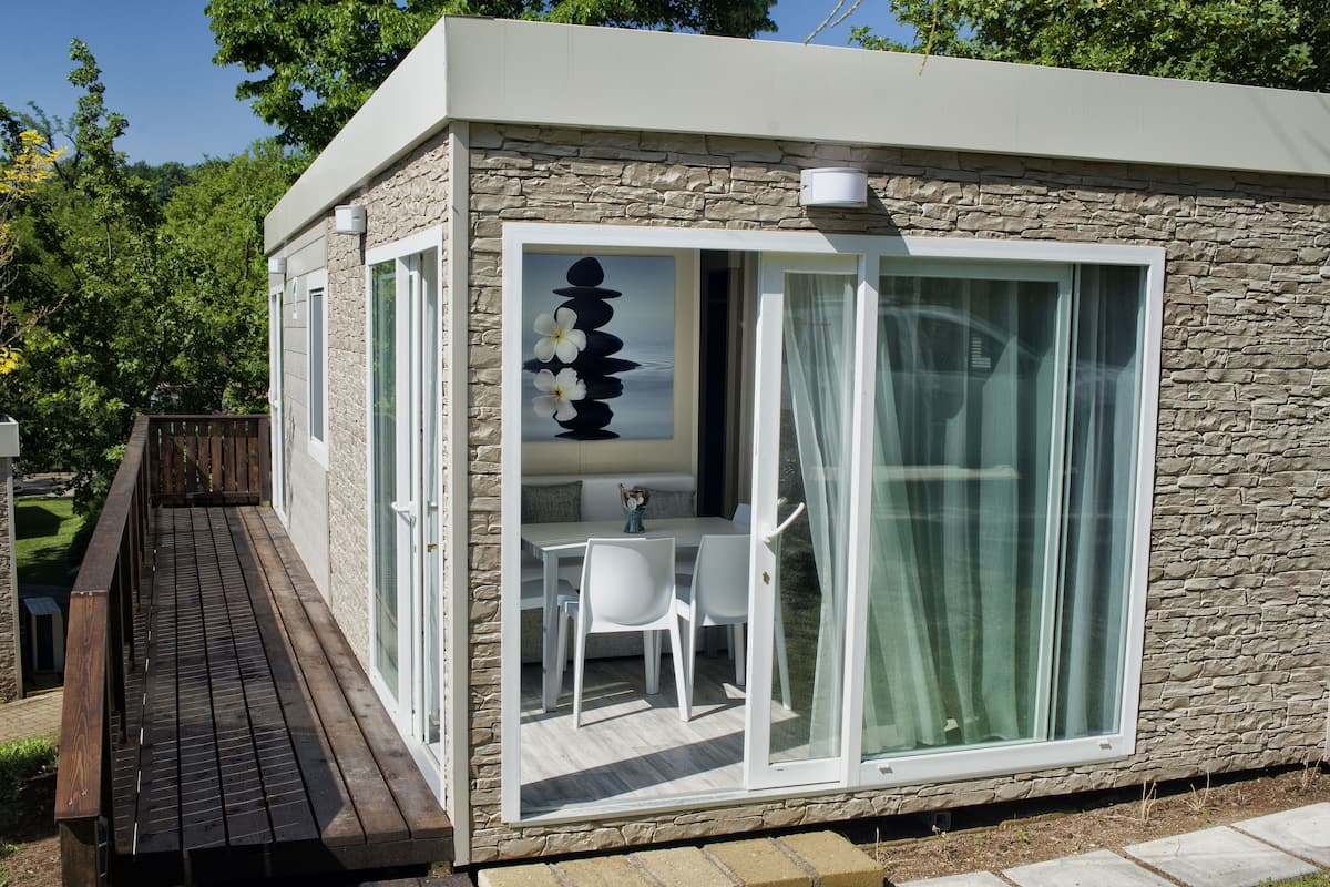 A modern building with a stone facade, a wooden deck, and a glass door leading to a dining area with white furniture.