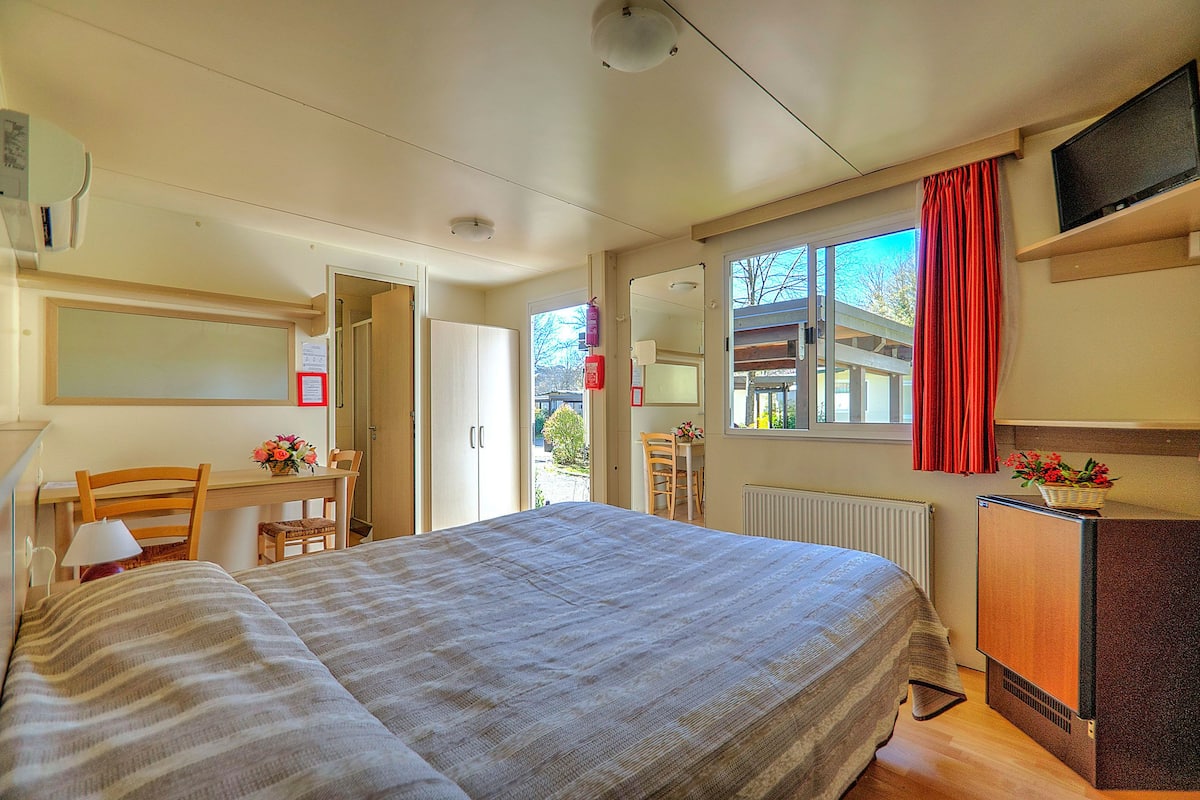 A bedroom with a bed, a desk, a chair, a dresser, a mirror, and a window with a red curtain.