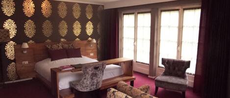 Habitación de hotel con una cama grande, cabecera de madera, sofá, sillón y ventana con cortinas.