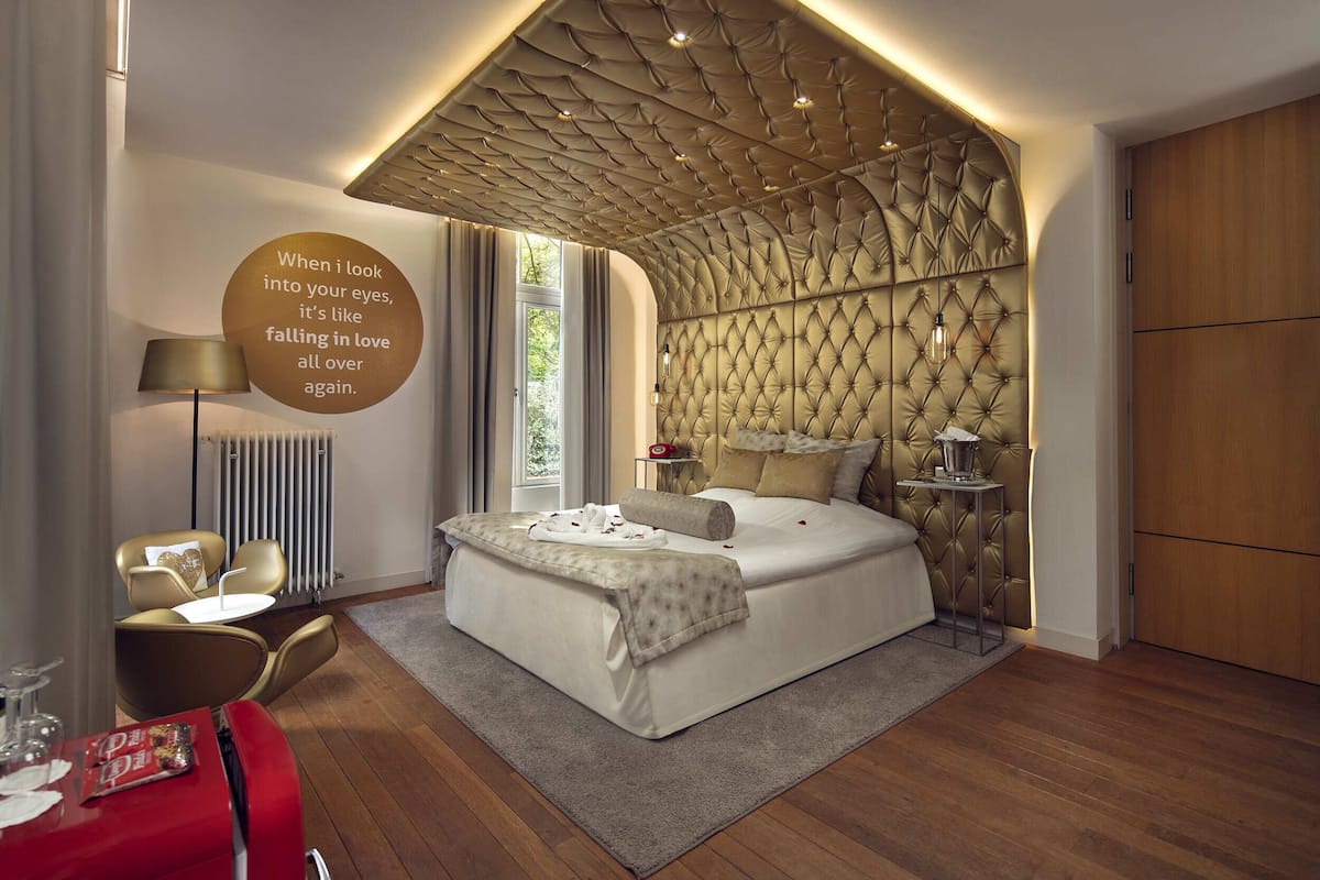 A modern bedroom with a tufted headboard, a bedside table with a lamp, and a red table with glasses.