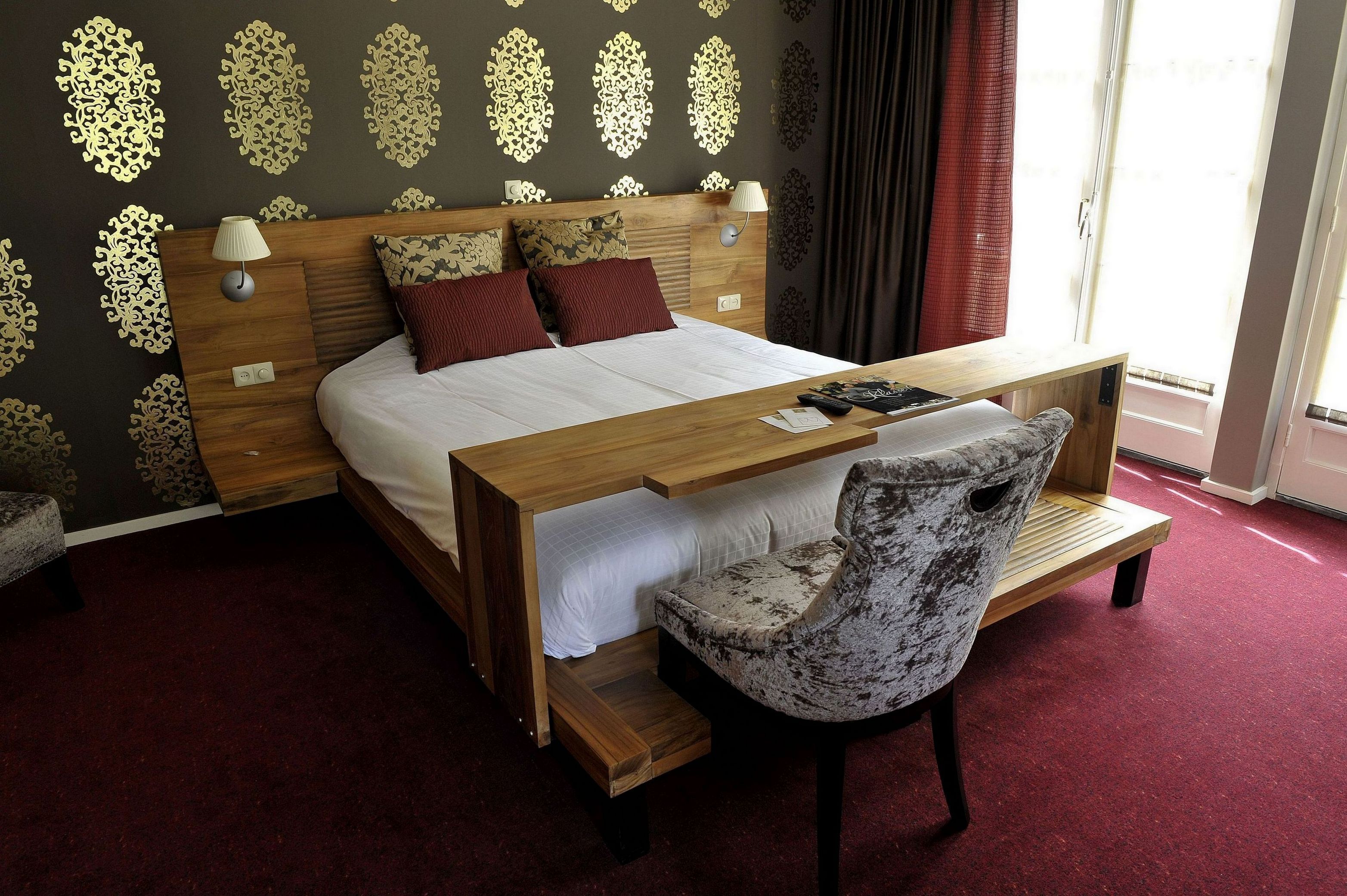 A bedroom with a wooden bed, two pillows, a bedside table with a lamp, and a chair.