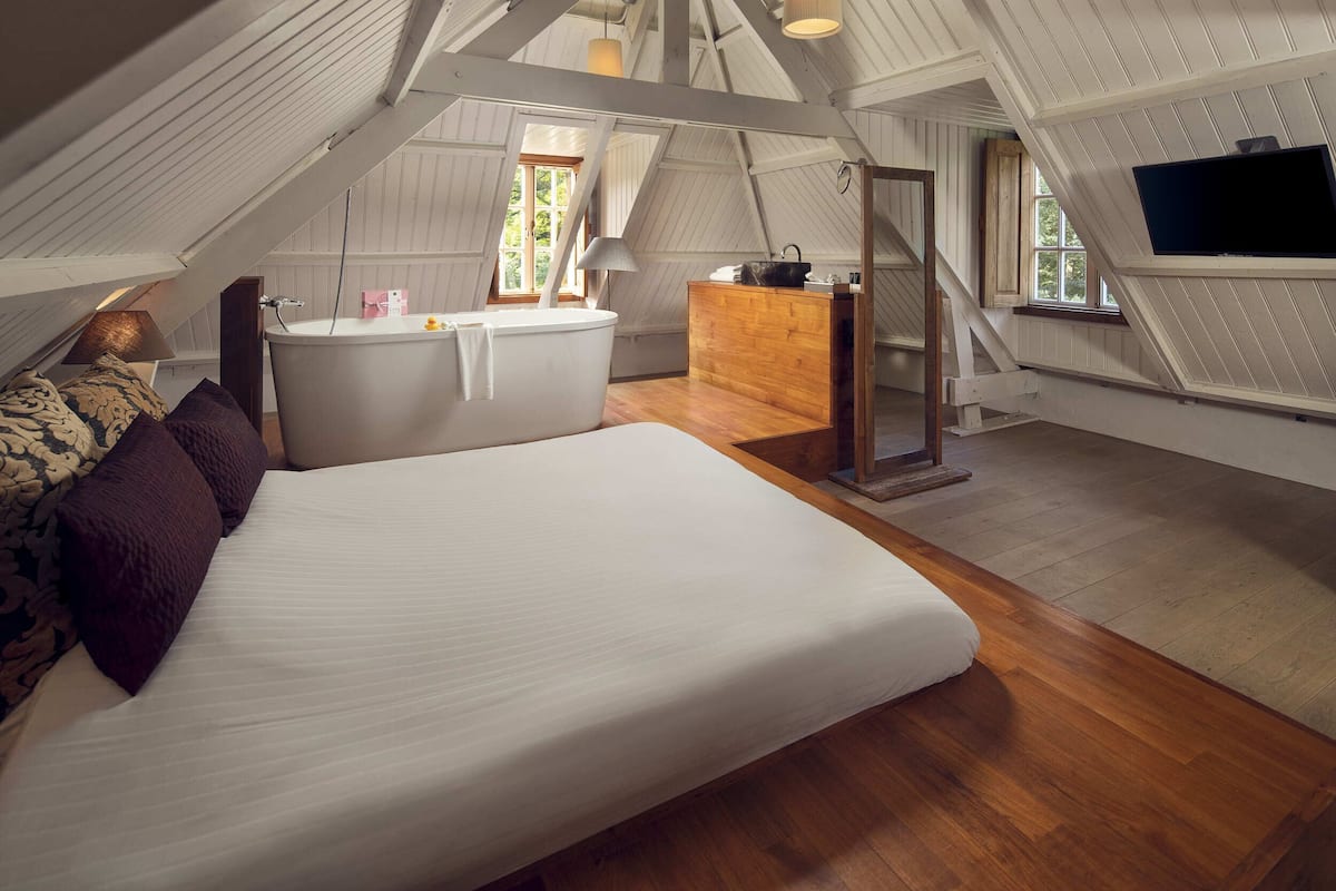 A bedroom with a large bed, a wooden dresser, a sink, and a television in a loft-style room with sloped ceilings and wooden floors.