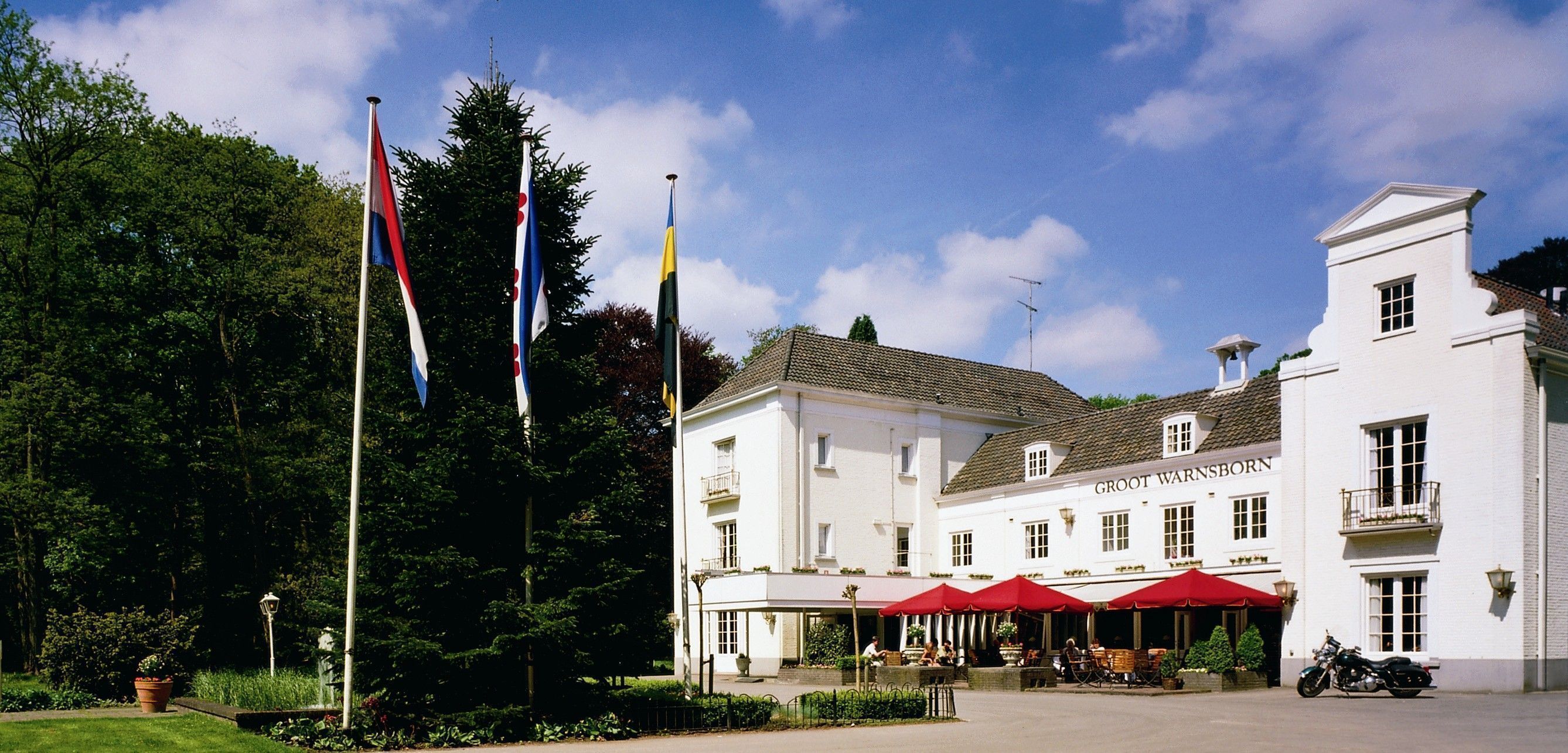 Ein weißes Gebäude mit einem Schild, auf dem „Groot Warnsborn“ steht, und einem Sitzbereich im Freien mit roten Sonnenschirmen.