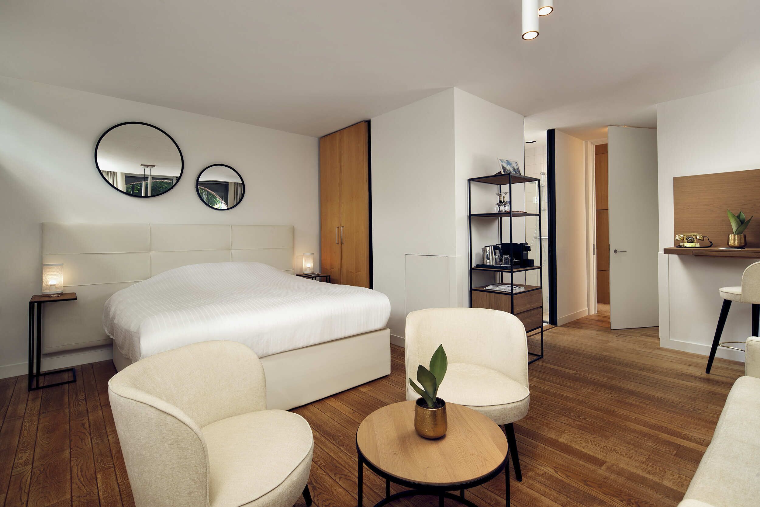 Een moderne hotelkamer met een groot bed, twee fauteuils, een kleine tafel met een plant, een bedlamp, een spiegel, een houten garderobe, een boekenplank en een bureau met stoel.