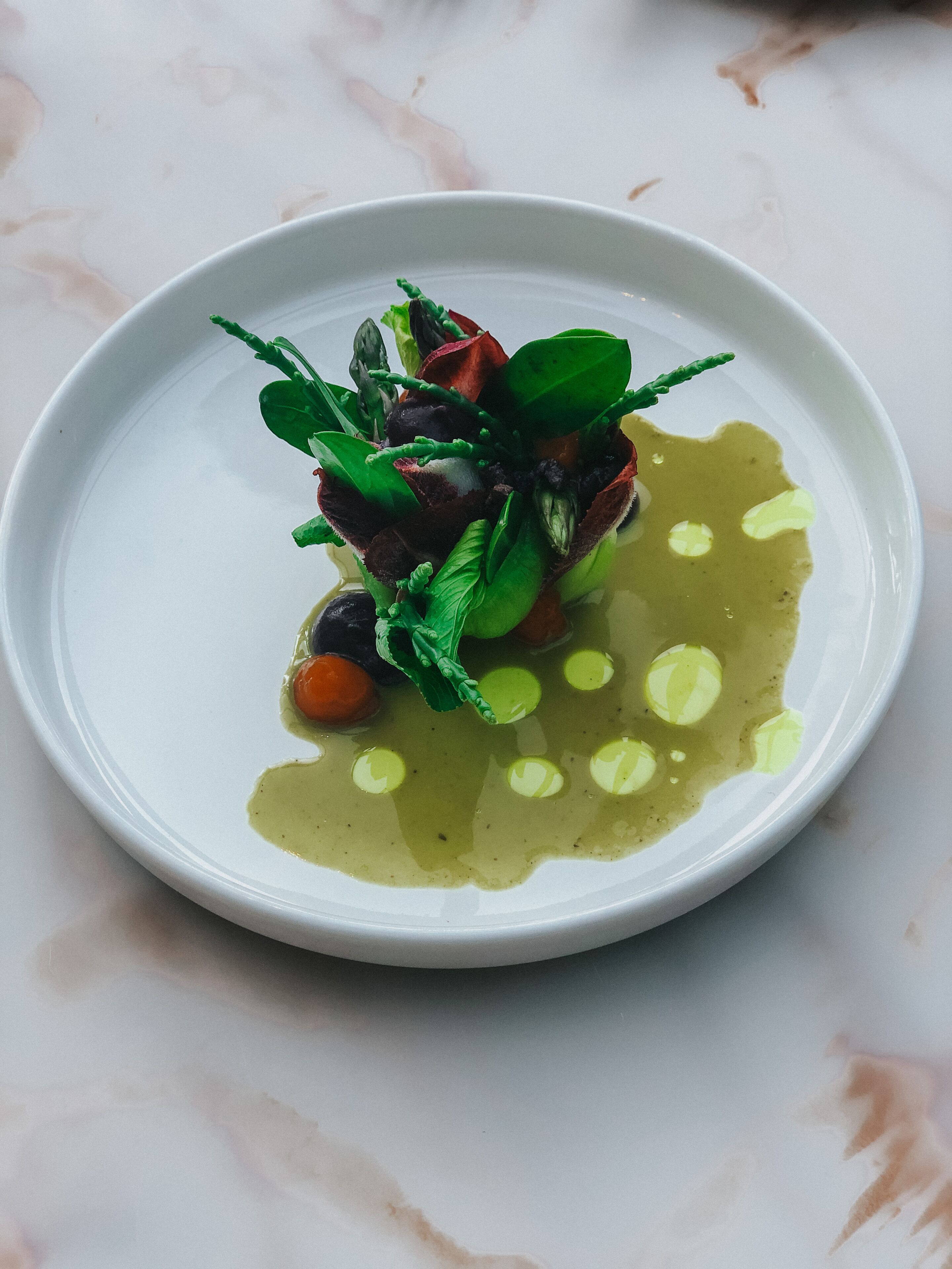 A white plate with a dish of food, including greens, a red element, and a yellow sauce.