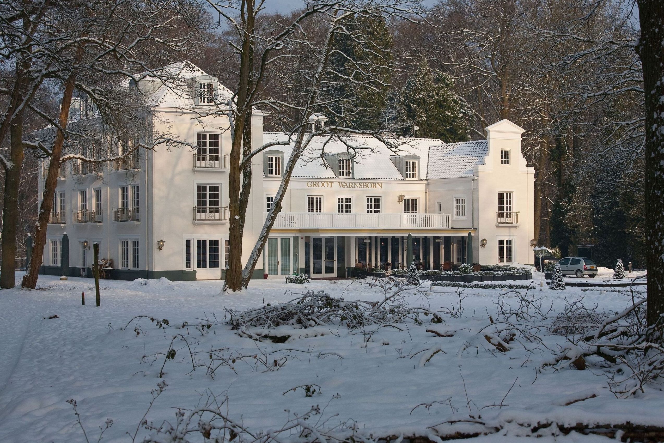 A large, white building with the name "Groot Warnsborn" on the facade, surrounded by snow-covered trees and a clear sky.
