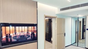 Quarto de hotel com uma televisão de tela plana grande fixada em um painel de madeira, porta deslizante que dá para um banheiro e um separador de vidro dividindo o quarto de outro espaço.