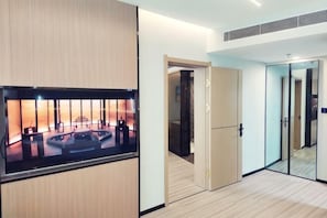 A hotel room with a large flat-screen TV mounted on a wooden panel, a sliding door leading to a bathroom, and a glass partition separating the room from another space.