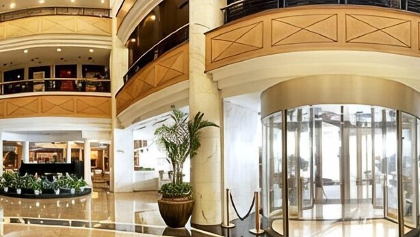 Um saguão de hotel com piso de mármore, um balcão de recepção, uma grande planta em vaso e um elevador de vidro.