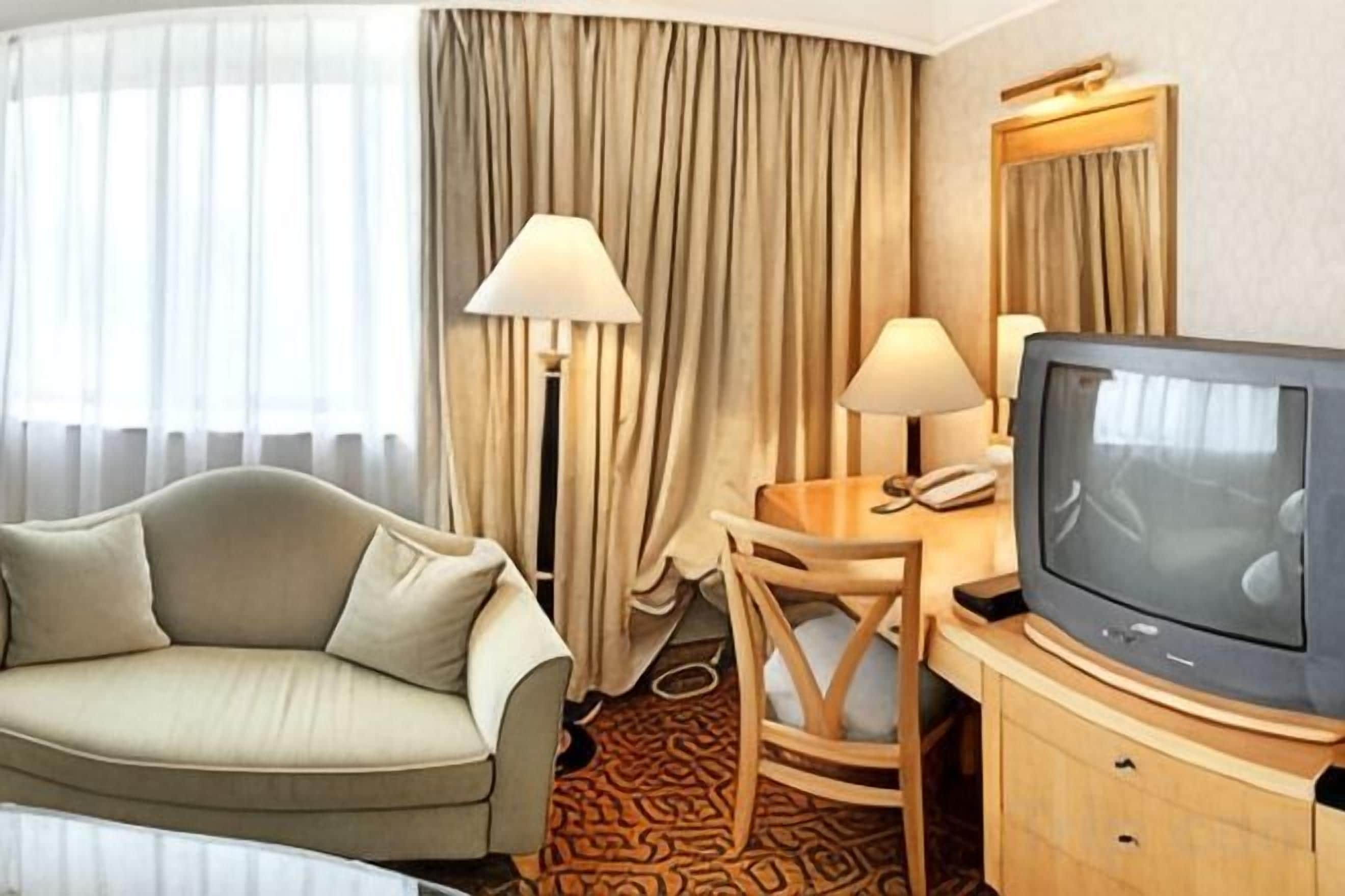 Una camera d'albergo con un divano, una scrivania con una lampada, una televisione e un'ampia finestra con tende.