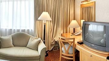 Una camera d'albergo con un divano, una scrivania con una lampada, una televisione e un'ampia finestra con tende.