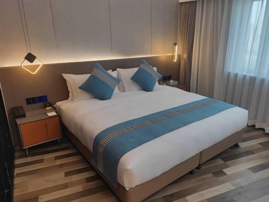 Habitación de hotel con una cama grande, una mesita de noche con teléfono y un panel de control en la pared, además de una ventana con cortinas.
