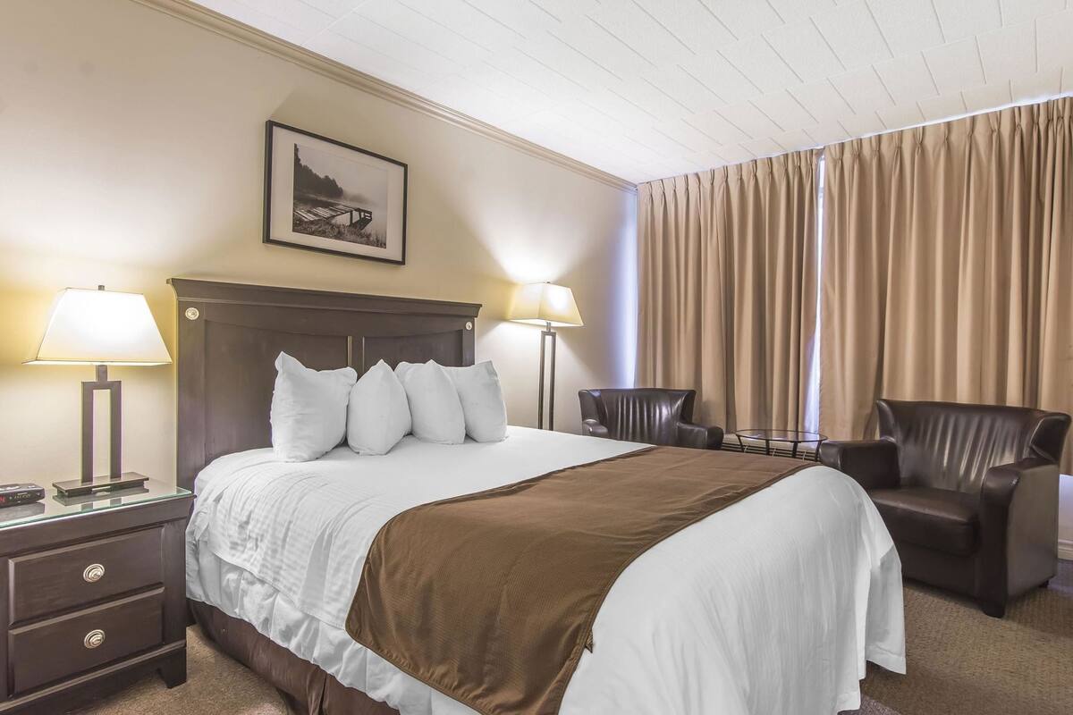 Una habitación de hotel con una cama, dos sillas, una mesita de noche con lámpara, una mesita auxiliar y un cuadro en la pared.
