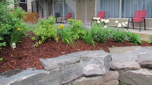 A landscaped garden with various plants and flowers, a stone pathway, and a building with numbered doors in the background. - Rodeway Inn King William (Huntsville)