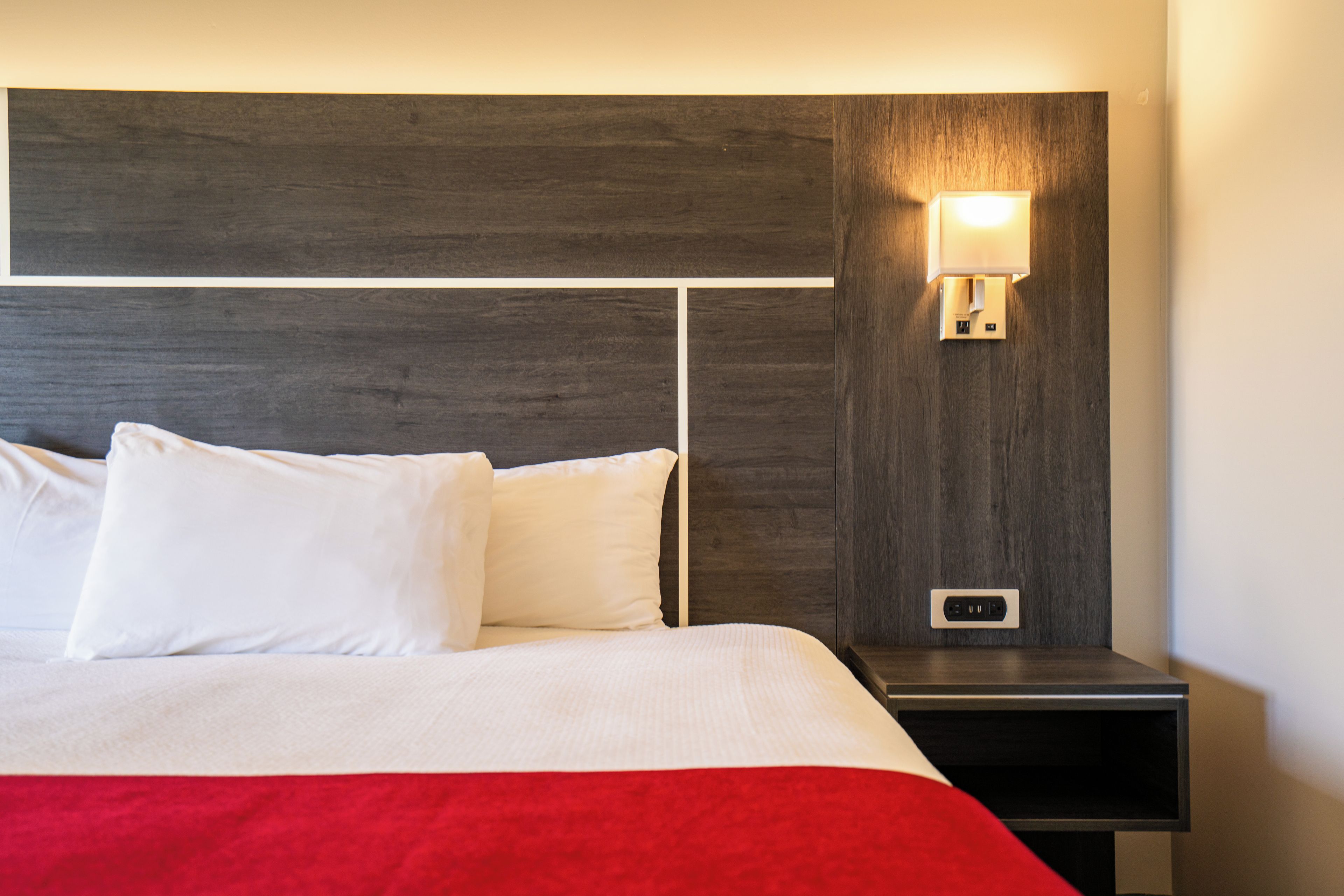 Una cama bien hecha con sábanas blancas y un detalle rojo, una mesita de noche con una lámpara de pared, y un enchufe.