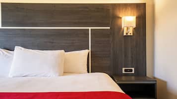 Una cama bien hecha con sábanas blancas y un detalle rojo, una mesita de noche con una lámpara de pared, y un enchufe.