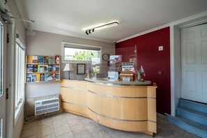 A reception desk with a wooden counter, a window with a view of greenery, a wall-mounted clock, and a sign indicating no cash accepted. - Rodeway Inn King William (Huntsville)