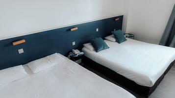 Two single beds with white linens and teal pillows, a bedside table with a phone, and a wall-mounted light switch.