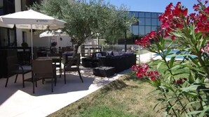 An outdoor seating area with tables and chairs, a white umbrella, and a pool in the background. - Aéroport Hôtel (Mauguio)