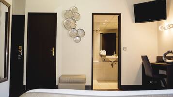 Habitación de hotel con cama, escritorio, silla, televisor montado en la pared y baño visible a través de una puerta abierta.